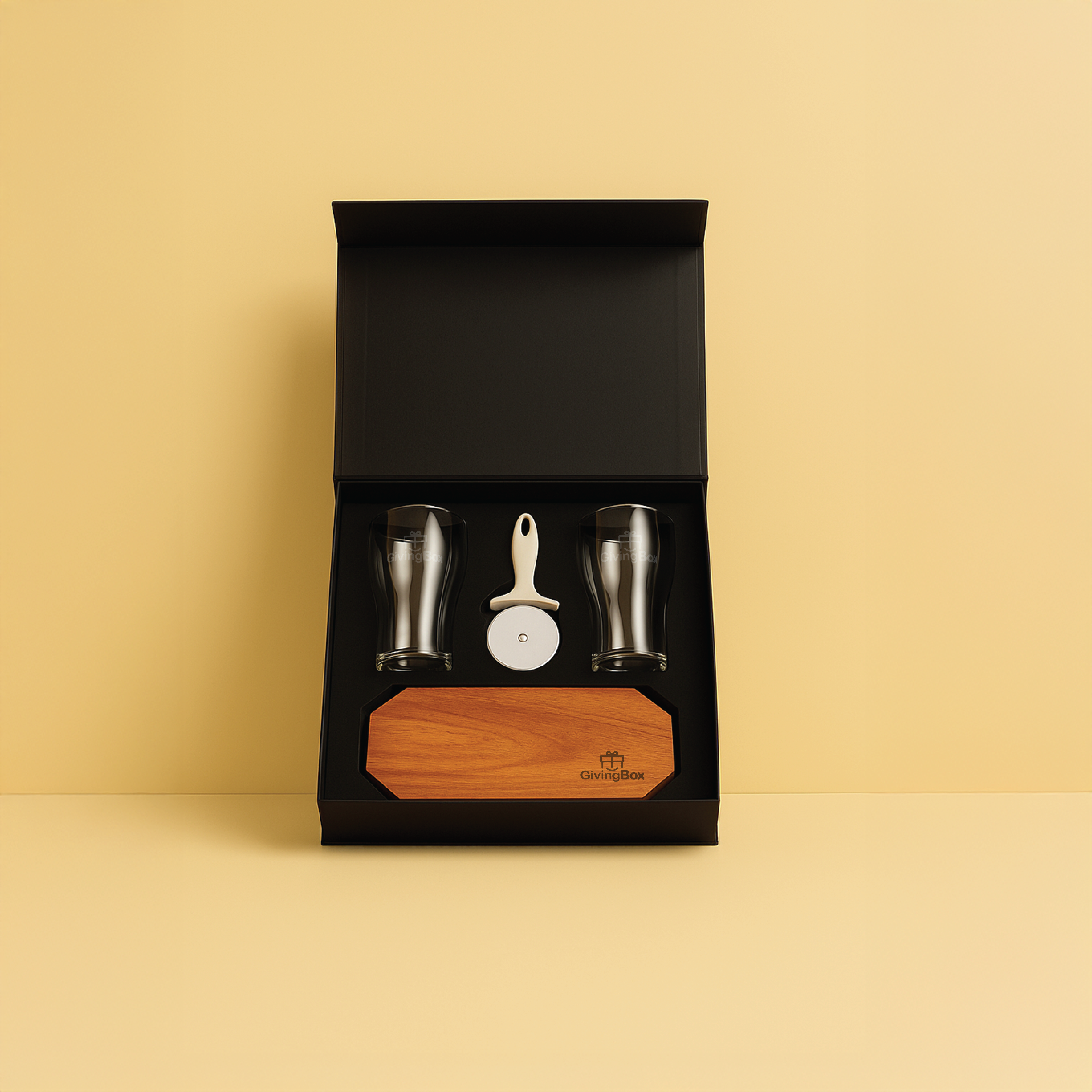Set pizzero con caja negra, dos vasos cerveceros, cortador de pizza y tabla de madera octogonal, sobre fondo amarillo.